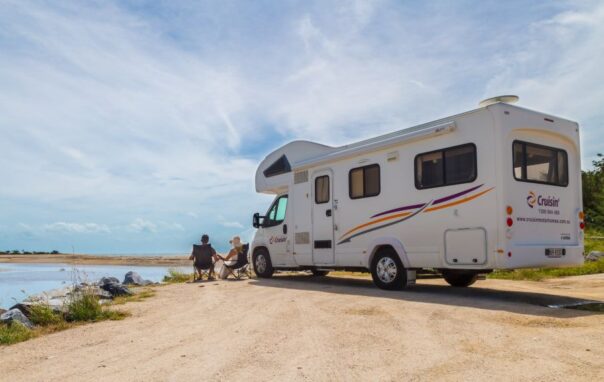 Cruisin' Discovery camper staat geparkeerd op het zand aan een meer, stel zit op campingstoelen buiten