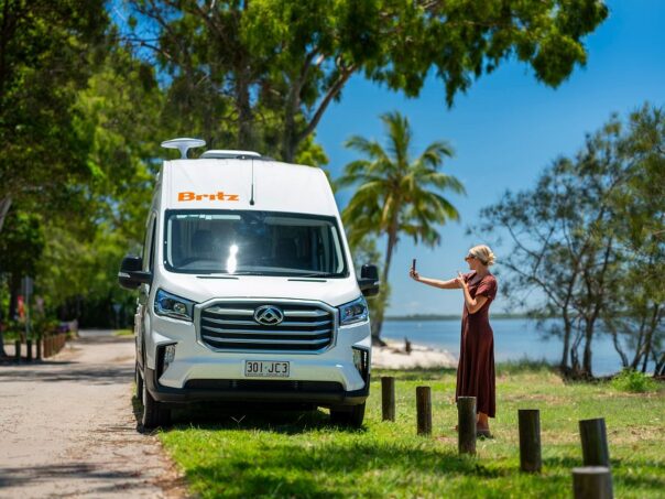 Vrouw maakt selfie naast de Britz Trailblazer in Queensland die bij het strand geparkeerd staat tussen palmbomen