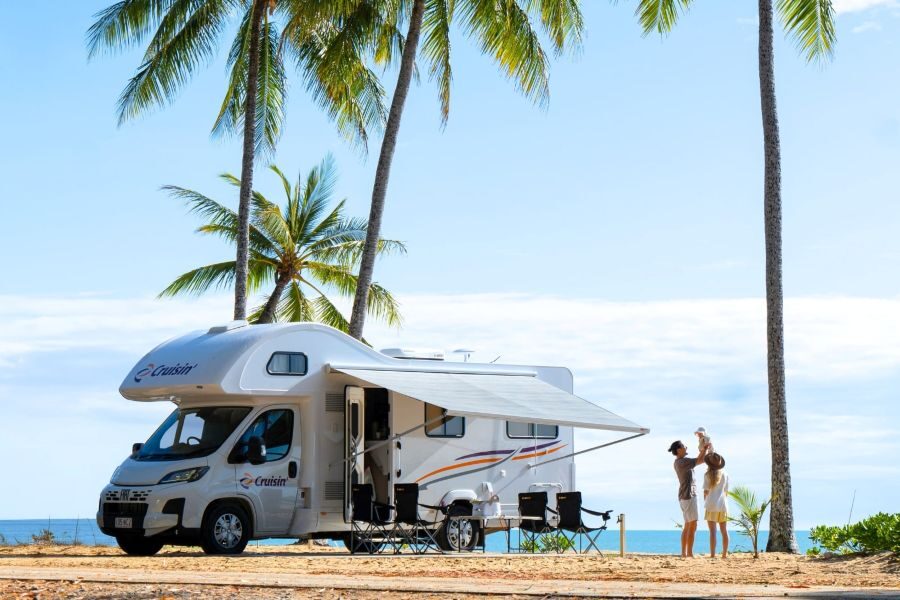 Vader tilt jong kind op naast Cruisin' 6 Berth Deluxe camper die tussen palmbomen aan het strand geparkeerd staat