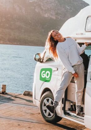 Een vrouw hangt uit de deur van de Go Cheap 4 Berth Hitop die geparkeerd is aan het water