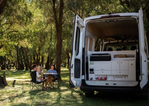 Jucy Coaster geparkeerd tussen bomen op een camping, achterdeuren zijn open en de keuken is te zien, stel zit buiten aan tafel