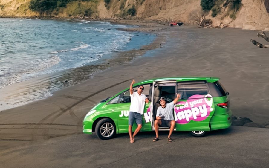 Jucy Crib staat geparkeerd op een strand, twee mannen zwaaien vanuit de zijdeur