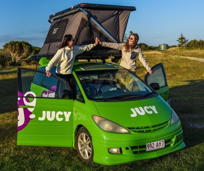 Jucy Crib Plus staat geparkeerd op het gras met de daktent open, twee vrouwen hangen uit de deuren en geven elkaar een high five