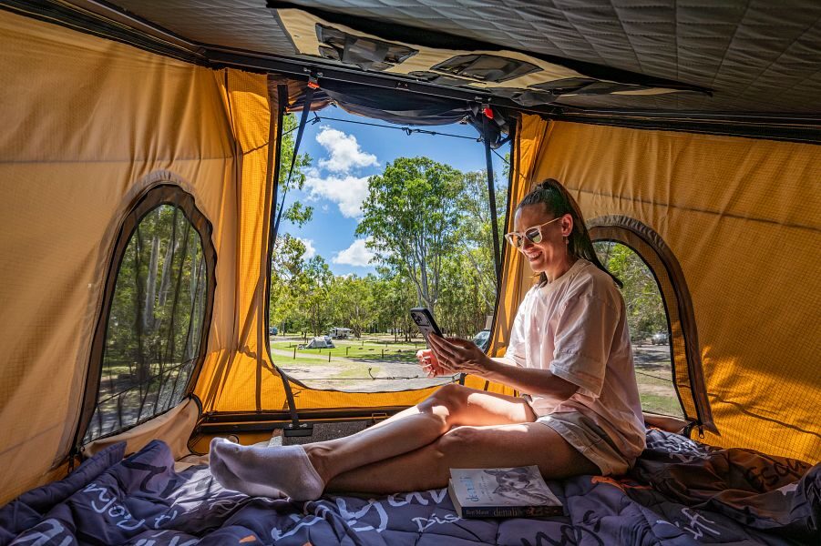 Britz Voyager Australië binnenkant van de pop up daktent met vrouw kijkend op haar telefoon en uitzicht op de camping