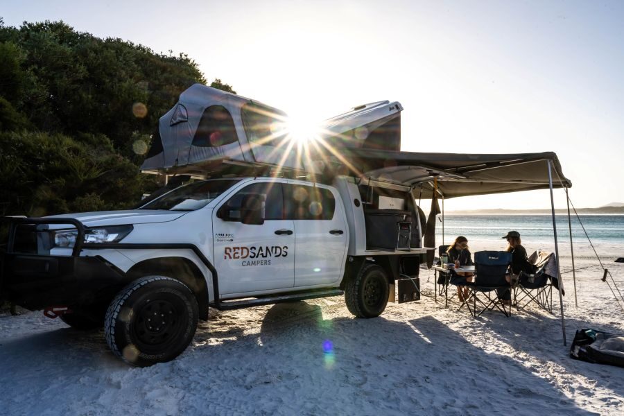 Twee camperreizigers genieten op het strand met hun gehuurde RedSands 4WD camper, zittend in hun campingstoelen onder de royale luifel. De zon schijnt tussen de twee daktenten door.