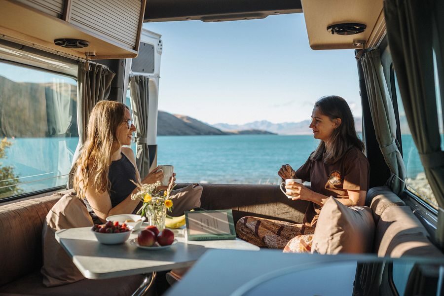 Twee vriendinen zitten achterin de Britz Venturer camper in de zithoek met wat te drinken en de achterdeuren open met uitzicht op een meer in Nieuw-Zeeland