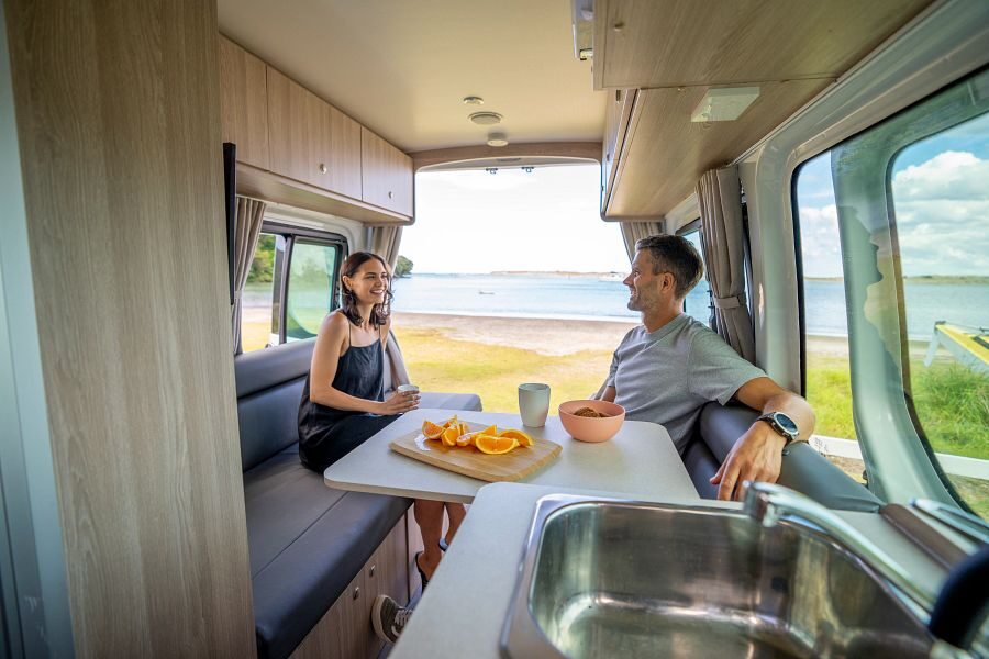 Een man en vrouw zitten in de zithoek met tafel aan de achterkant van de Britz Venturer camper met openstaande achterdeuren en uitzicht op het water in Nieuw-Zeeland
