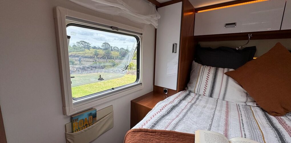 Het tweepersoonsbed achterin de Britz Odyssey camper met kasten boven het bed en een raam met uitzicht op de natuur van Nieuw-Zeeland