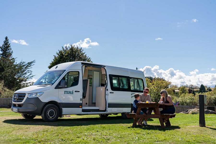 Een gezin zit aan een picknicktafel voor de Maui Ultima Plus camper die geparkeerd staat op het gras