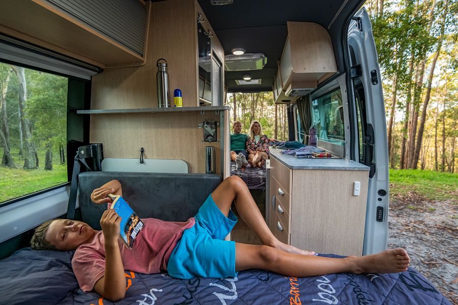 Een kind ligt voorin de camper op het eenpersoonsbed een boek te lezen terwijl de ouders achterin de camper op het tweepersoonsbed zitten