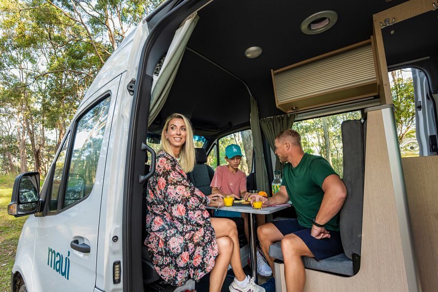 Een man en een vrouw met een kindje zitten voorin de Maui Ultima Plus camper in de zithoek aan tafel een glas jus d'orange te drinken