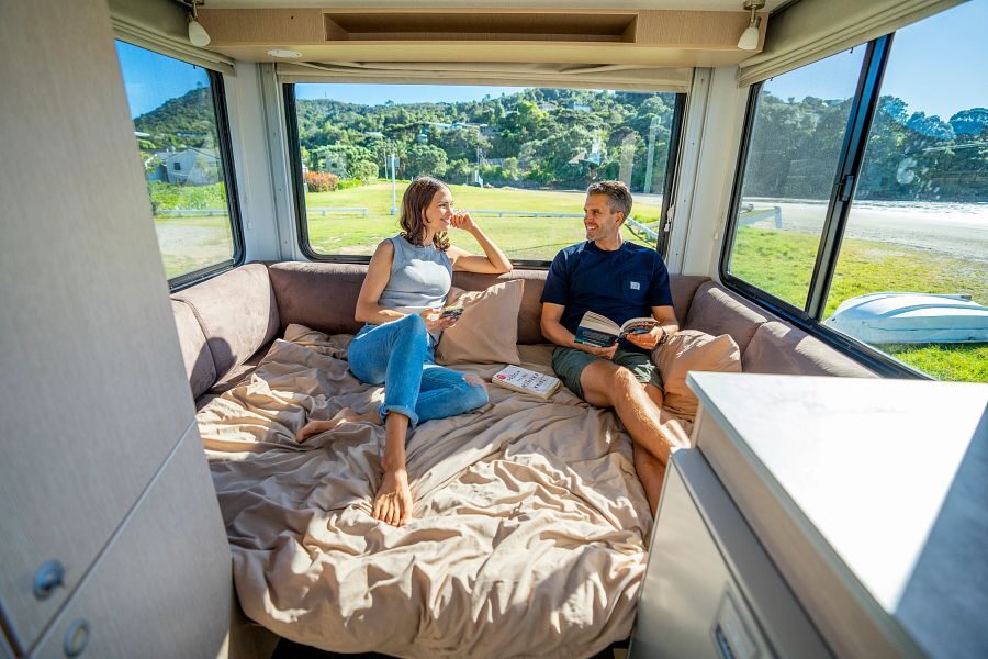 Een koppel zit achterin de Maui Cascade camper op het tweepersoonsbed met aan iedere kant ramen en uitzicht op de natuur van Nieuw-Zeeland