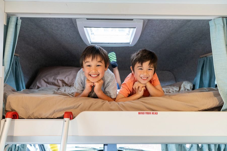 Twee kinderen liggen lachend in het bed in de alkoof boven de bestuurdersruimte van de Maui Beach camper