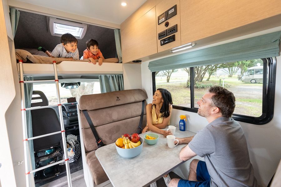 Twee kinderen liggen in het tweepersoonsbed in de alkoof boven de bestuurdersruimte die bereikbaar is via een ladder en de ouders kijken lachend naar de kinderen vanuit de zithoek in het midden van de Maui Beach camper