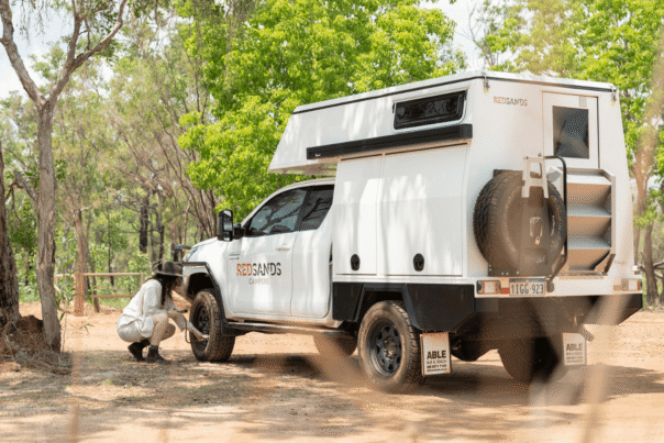 De camperreizigster is bezig met het meten van de bandenspanning van de Redsands camper. Aan de achterzijde van de camper is een reservewiel bevestigd en een opklapbaar trapje naar de achterdeur