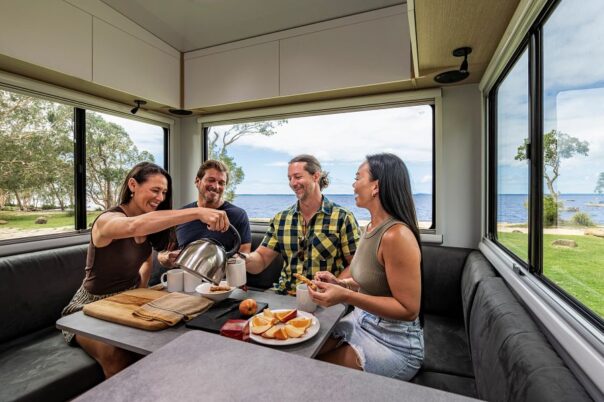 Vrienden zitten achterin de zithoek van de Maui River camper te eten en te drinken met vanuit de grote ramen uitzicht op het water en de natuur