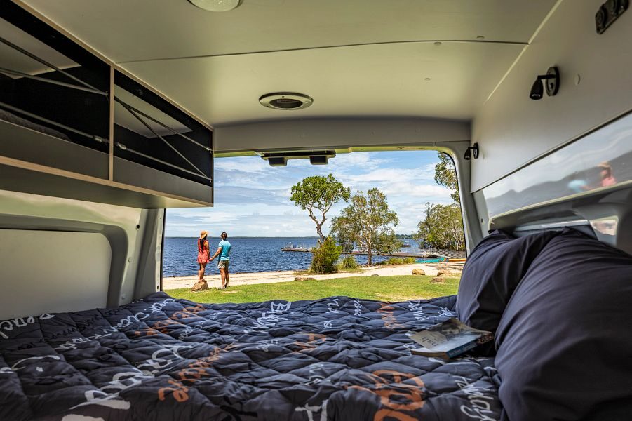 Het tweepersoonsbed in de Apollo Hitop camper in Australië met de achterdeuren open waar een koppel staat te genieten van het uitzicht op het water