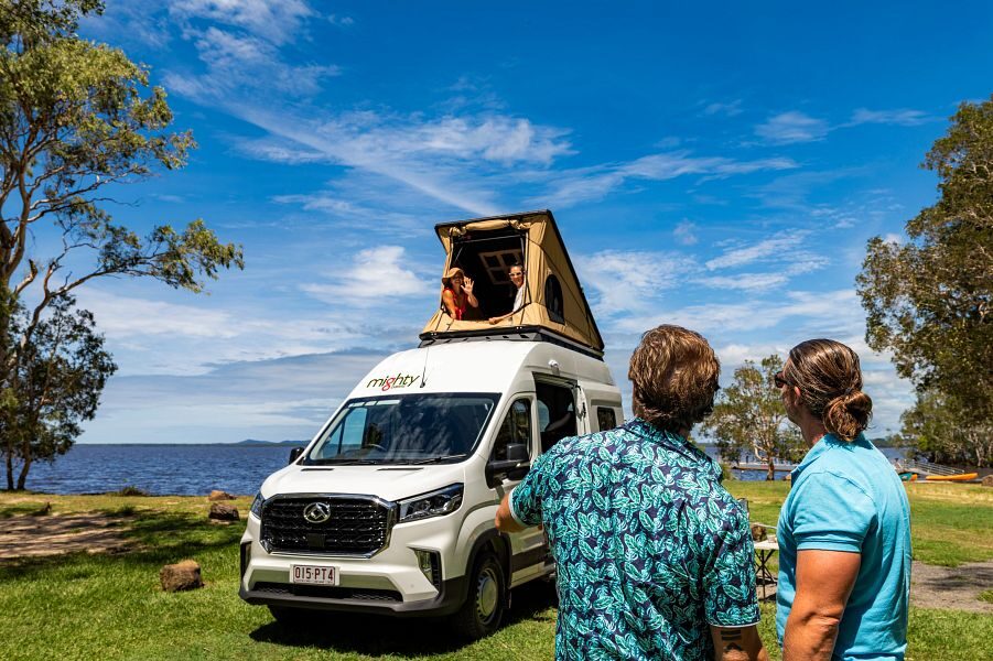 Twee mannen kijken naar twee vrouwen die bovenin de daktent zitten op de Mighty Double Down camper die te huur is in Australië en Nieuw-Zeeland