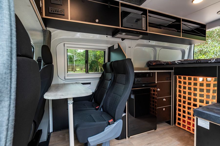 Het interieur van de Mighty Double Down camper met achter de bestuurdersruimte twee stoelen, een tafel, de keuken en een tweepersoonsbed