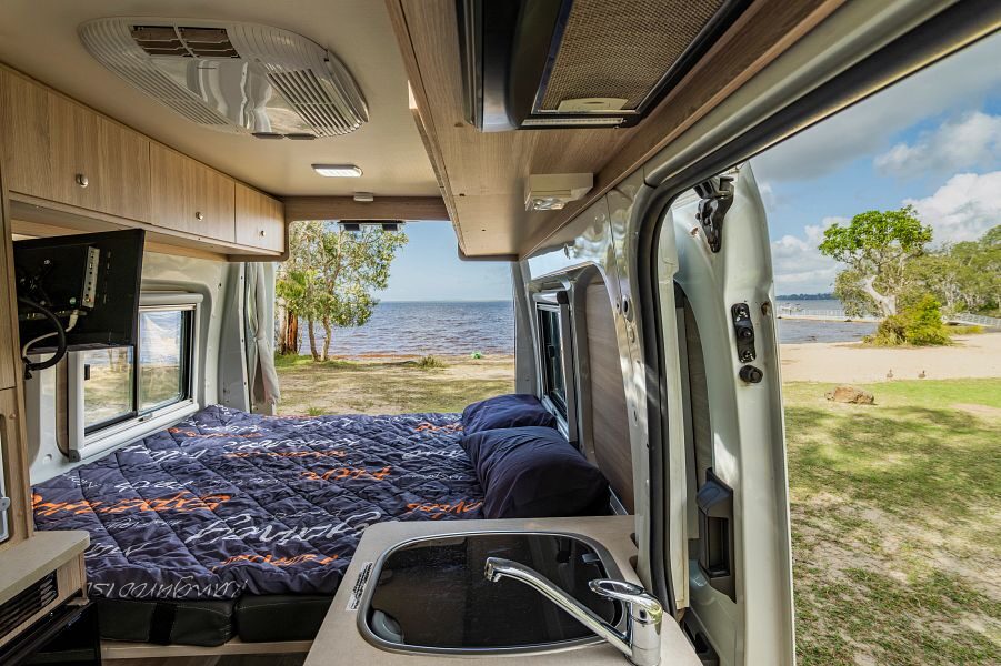 Het interieur met tweepersoonsbed van de Mighty Duo camper die geparkeerd staat op het gras met uitzicht op zee
