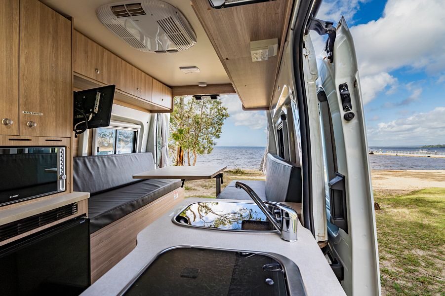 Het interieur met zithoek die je kunt ombouwen tot tweepersoonsbed van de Mighty Duo camper die geparkeerd staat op het gras met uitzicht op zee
