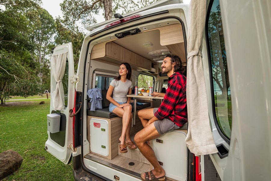 Een koppel zit aan tafel achterin de Mighty Duo camper met de achterdeuren open kijkend naar buiten