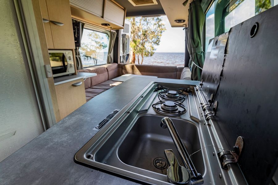 Het keukenblok met wastafel en twee gaspitten in de Mighty Deuce Plus camper met daarachter de zithoek en uitzicht op het water