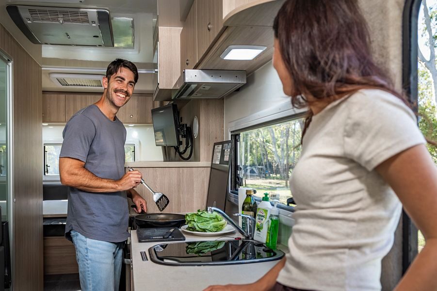 Een man is aan het koken en lacht naar zijn vrouw die in de deuropening van de Mighty Double Up camper staat