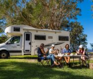 Twee stellen zitten buiten te kaarten met een Mighty camper op de achtergrond