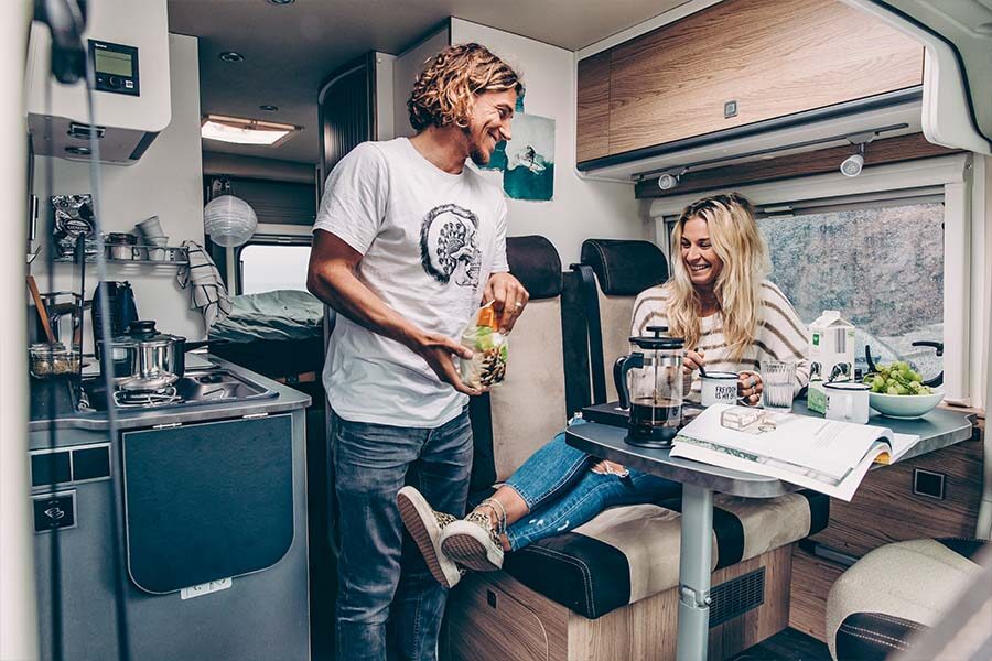 Een vrouw zit in het zitgedeelte aan tafel in de McRent Urban Plus camper terwijl haar man in de keuken staat en naar haar lacht
