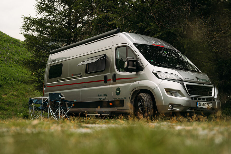 De McRent Urban Plus camper staat geparkeerd op een grasveld met campingstoelen aan de zijkant van de camper
