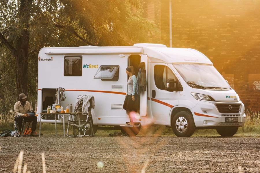 Het zij aanzicht van de McRent Kiwi Compact camper waar een vrouw op het trapje bij de ingang van de camper staat en een man voor de camper zit op een stoel