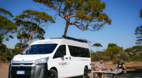 De camper is geparkeerd voor een tussenstop in een omgeving met bomen en twee personen zitten op hun campingstoelen en nuttigen een beker jus d'orange