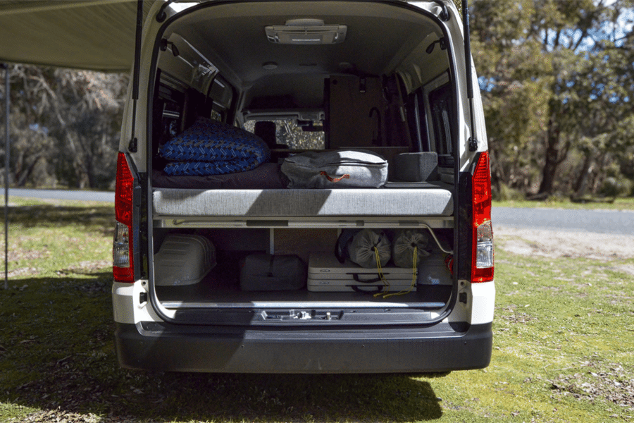 De achterzijde van de camper met de achterklep geopend waardoor het bed en de opbergruimte eronder zichtbaar zijn. De luifel is uitgezet.