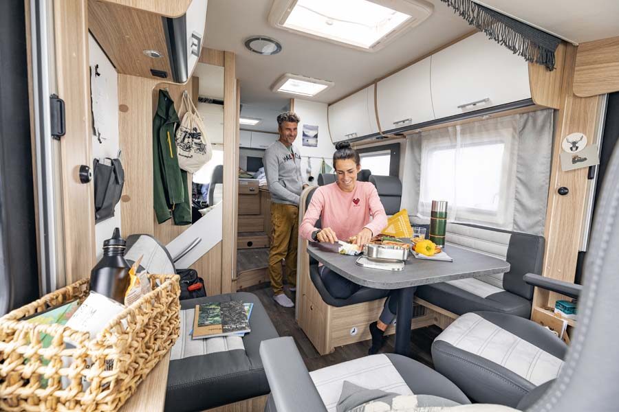 Een vrouw zit aan tafel in de McRent Comfort Luxury camper terwijl haar man in de keuken staat