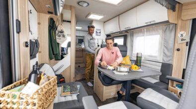 Een vrouw zit aan tafel in de McRent Comfort Luxury camper terwijl haar man in de keuken staat
