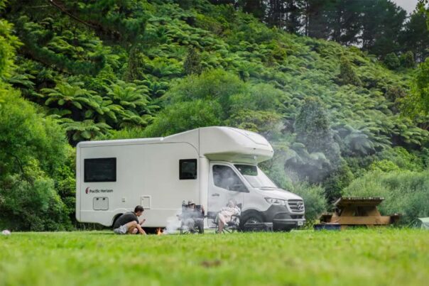 Een gezin zit voor de Pacific Horizon Gem 4 camper op de campingstoelen met een zelfgemaakt kampvuurtje in de groene natuur van Nieuw-Zeeland