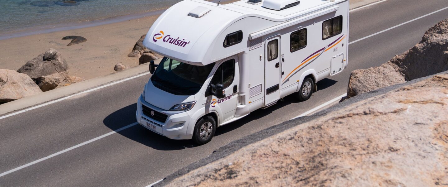 Cruisin' Seeker camper rijdt op een weg langs het strand waar iemand aan het suppen is in zee