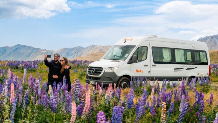 Stel maakt een selfie voor een Britz camper met lupines op de voorgrond