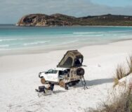 Een 4WD camper van Adventure rentals staat geparkeerd op het witte strand met een vrouw in een campingstoel ernaast